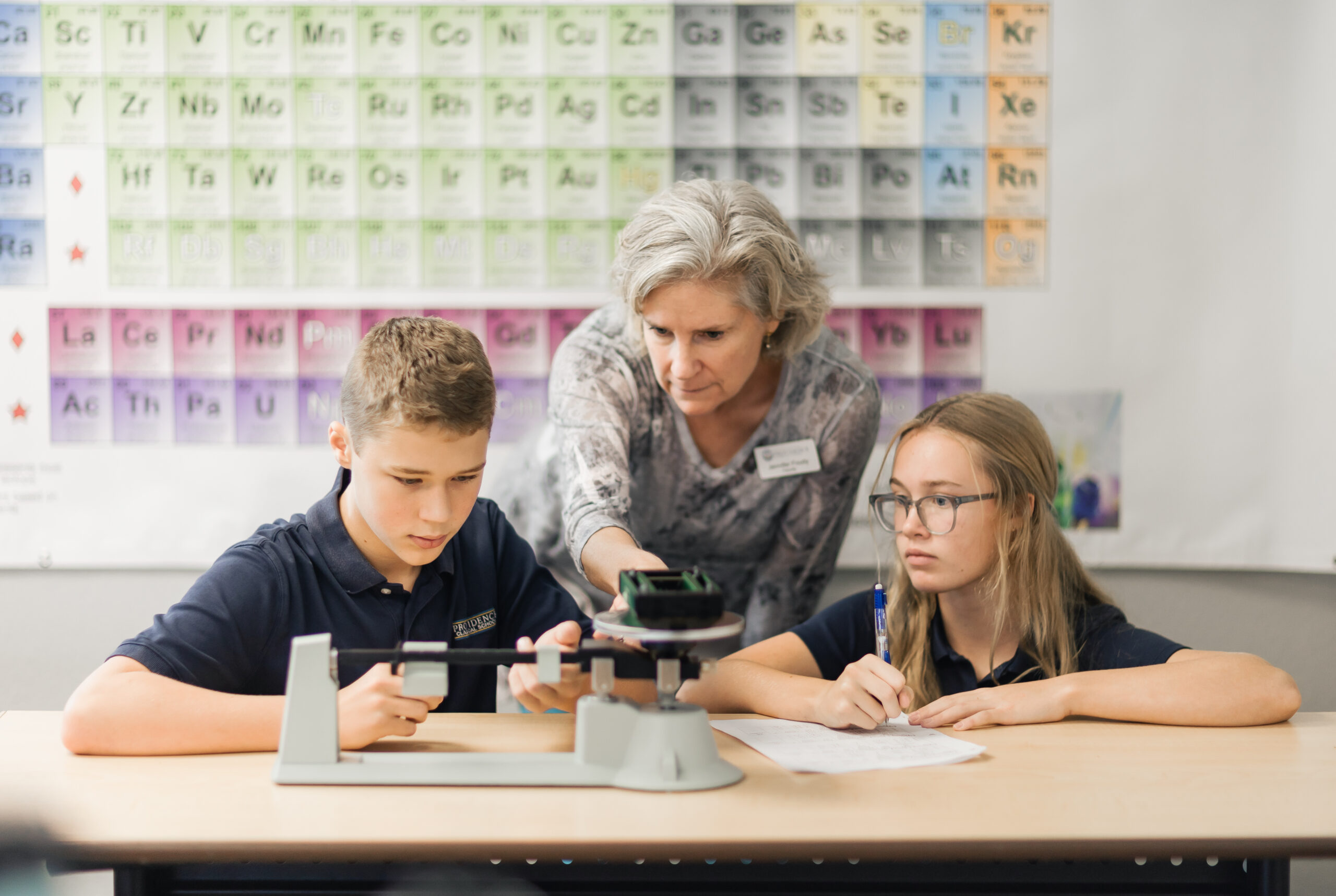 Physical Science students at Providence Classical School in Spring, TX, work with their teacher to measure the mass of an object using a scale.